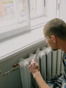 Handwerker misst mit dem Zollstock an einem alten gusseisernen Heizkörper unter einem Altbaufenster
