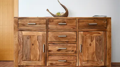 Massivholz-Sideboard mit drei Schubladen und zwei Türen vor heller Wand, warmes Licht auf der Maserung