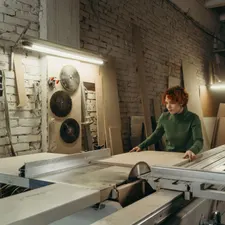 Tischlerin führt eine Holzplatte an der Formatkreissäge, Werkstatt mit Ziegelwand