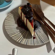 Angeschnittener Käsekuchen mit Kirschkompott auf gestreiftem Teller, daneben Gabel und Cappuccino im Sonnenlicht.