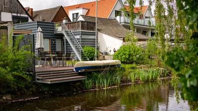 Blick vom Wasser auf das Haus mit Holzterrasse, Sonnenschirm und Kanu am Schilfufer