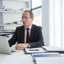 Steuerberater mit Brille im dunklen Anzug arbeitet konzentriert am Laptop, Aktenregale und Fenster im Büro